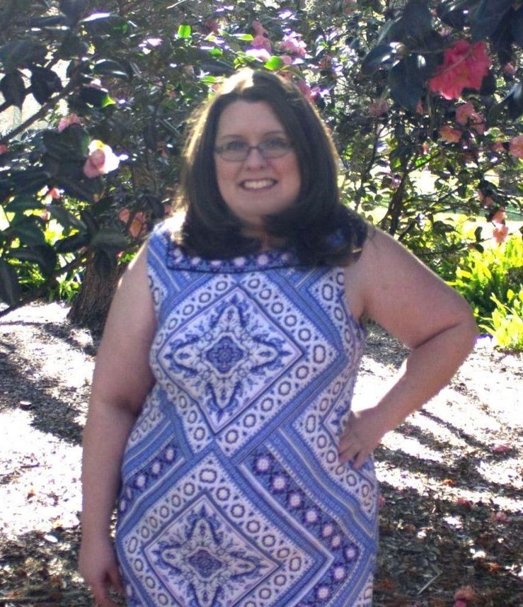 Woman with brown hair and glasses in a blue and white dress standing in front of a tree